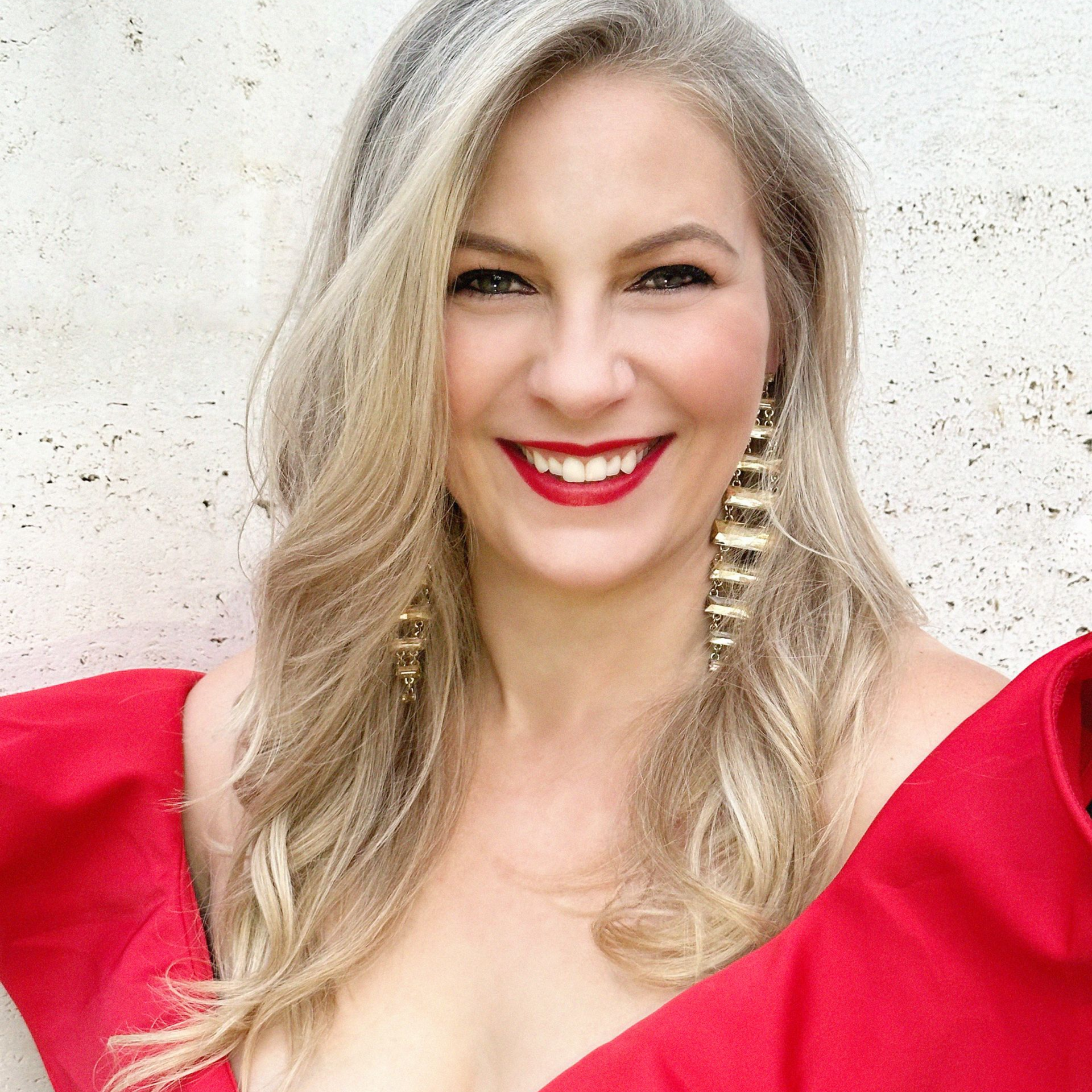 Woman with silver-blonde hair smiles, wearing a red dress with ruffled neckline and large gold earrings.