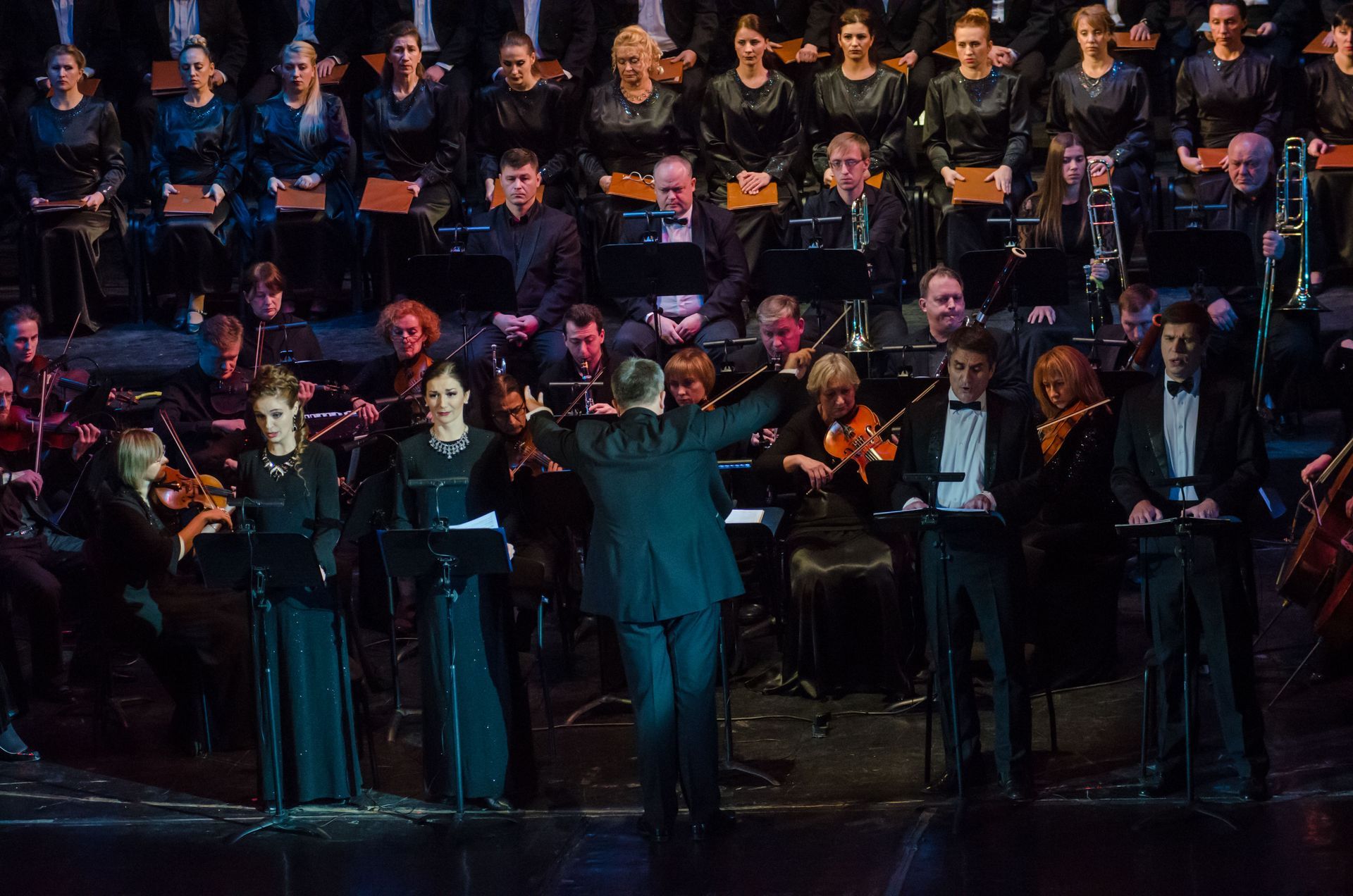 Conductor leading orchestra and choir in performance. Dark stage, formal attire, focused expressions.