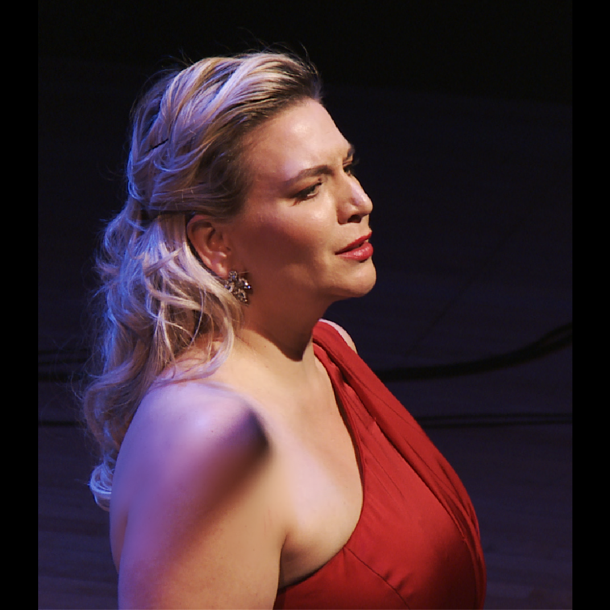 Woman with blonde hair in a red dress, singing onstage with a focused expression.