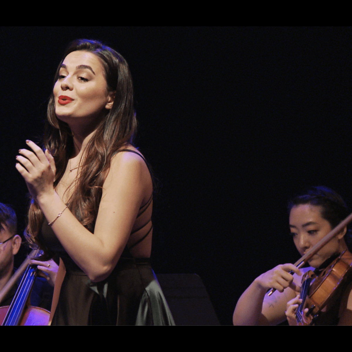 Woman singing on stage with musicians. She wears a dark dress, red lipstick.