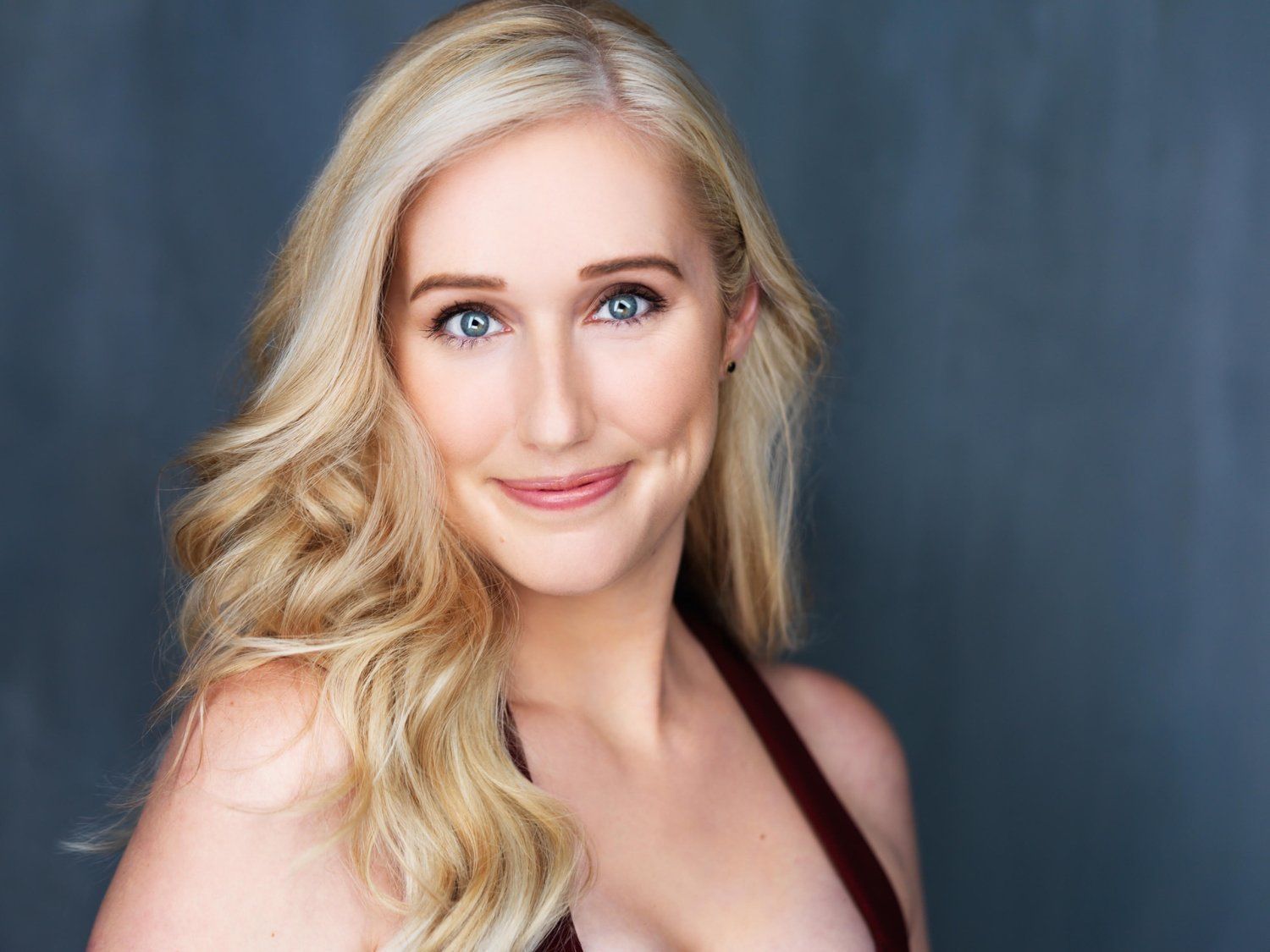 Blonde woman with blue eyes smiles softly, wearing a red halter top, against a gray background.