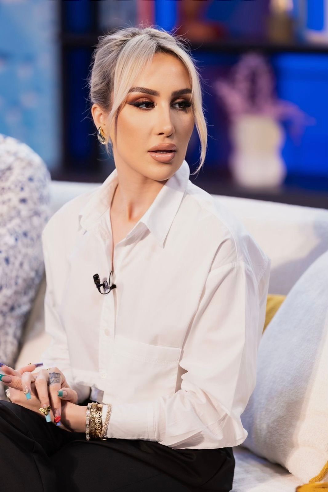 Woman with blonde hair in a white shirt on a set. She looks to the side, with jewelry and makeup.