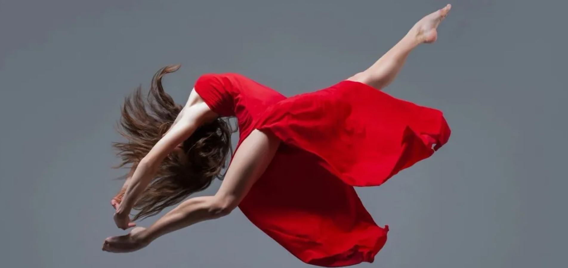 A dancer in a flowing red dress leaps, hair flying, against a gray background.