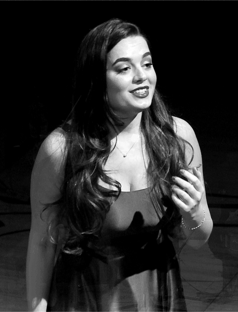 Woman with long hair, wearing a dress, smiling and gesturing onstage in black and white.