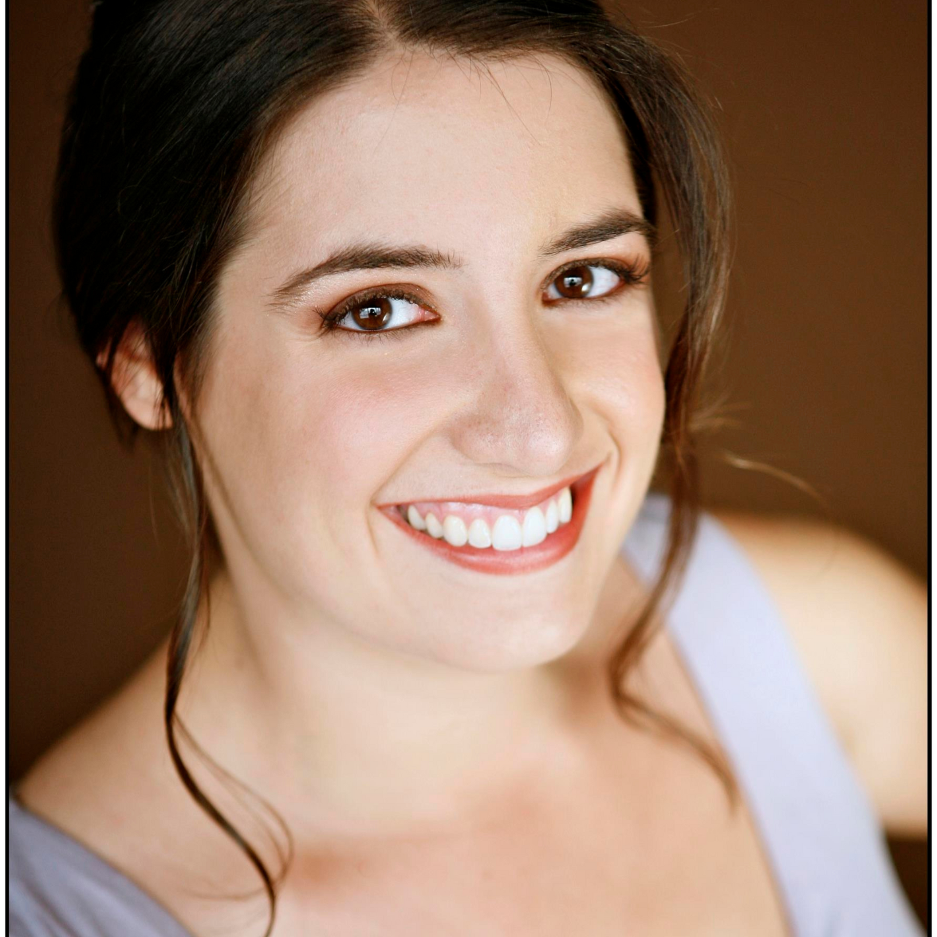 Woman with dark hair smiles at camera, wearing a light purple top, brown background.