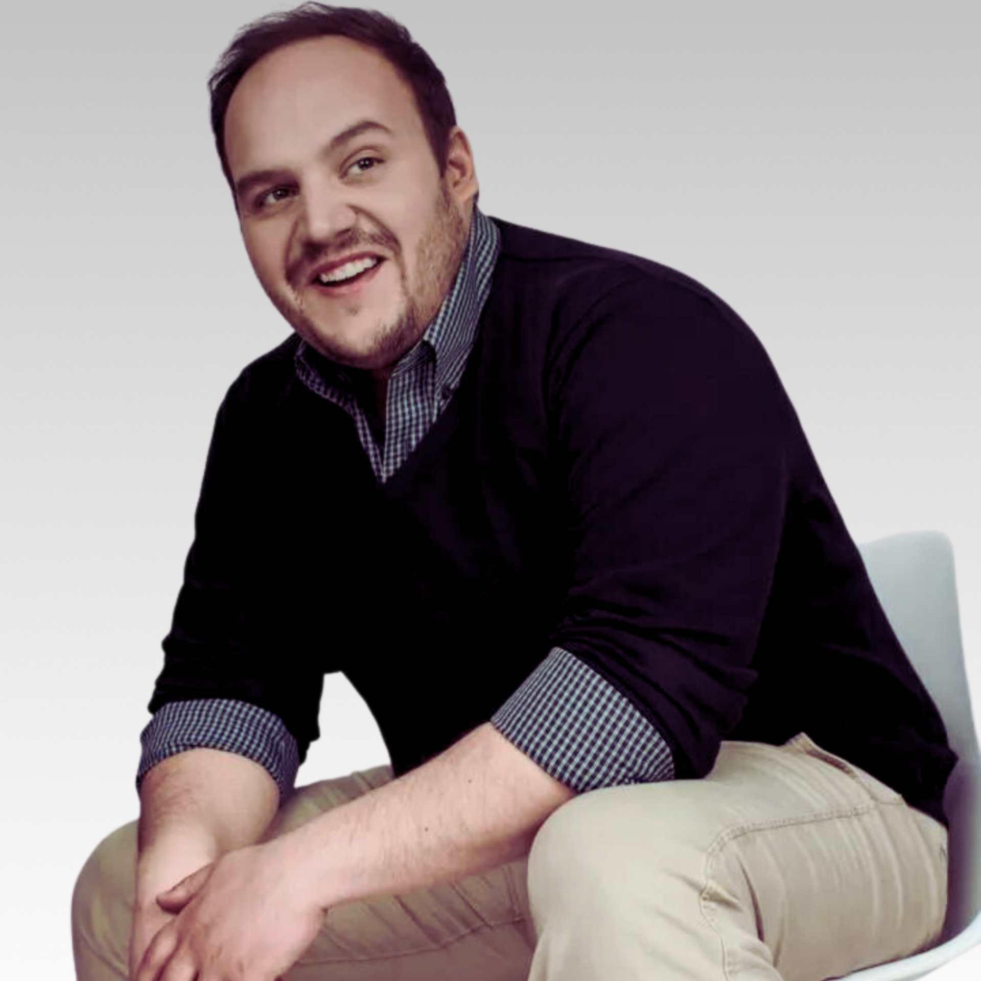Man sitting, smiling, wearing a dark sweater over a collared shirt and beige pants. White chair.