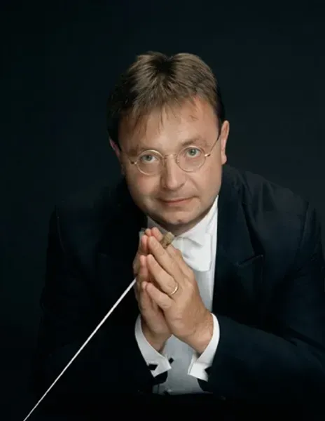 Man in tuxedo with baton, conducting orchestra, looking forward with glasses and serious expression.