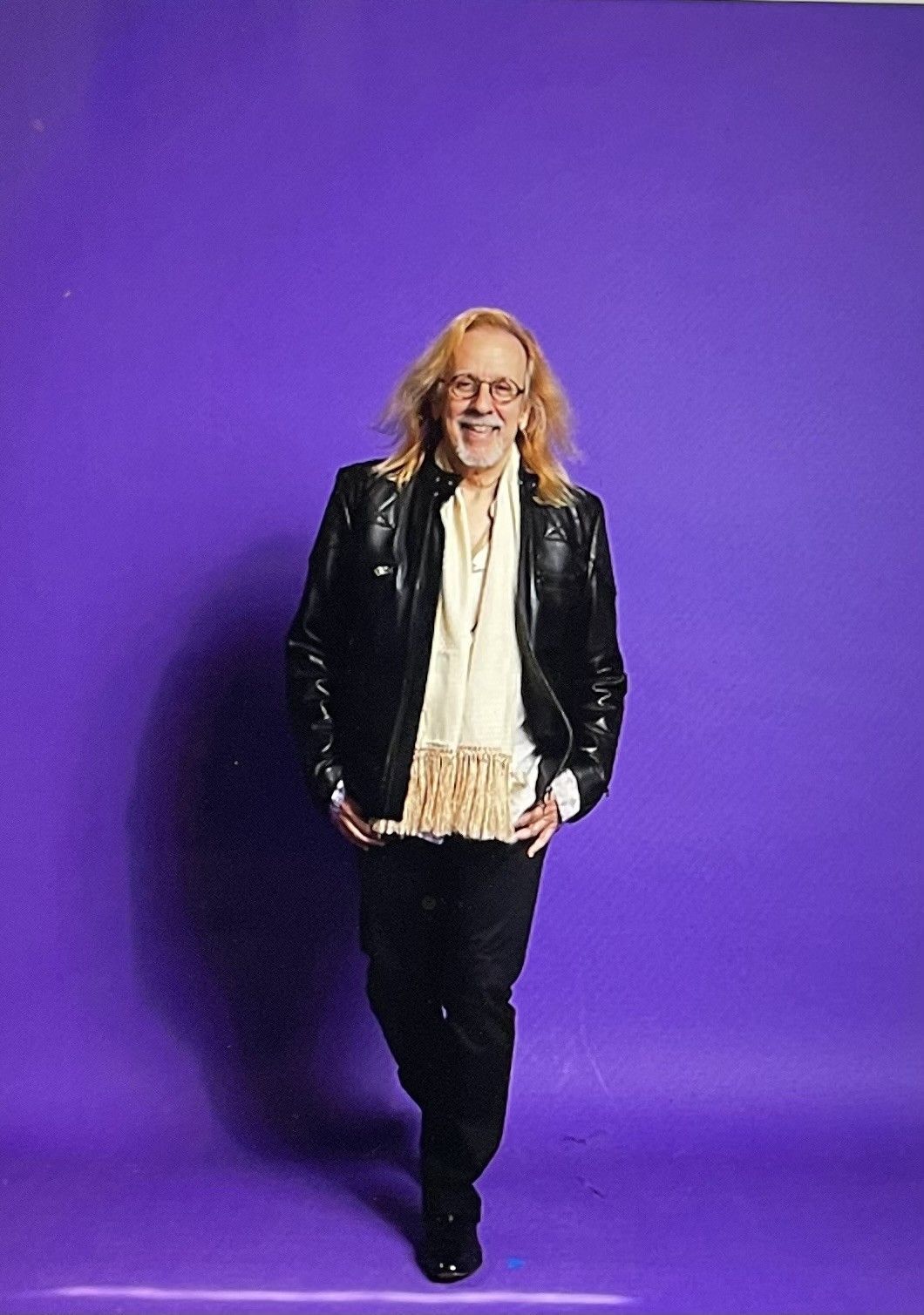 Man with long blond hair, leather jacket, and scarf, smiling against a purple backdrop.