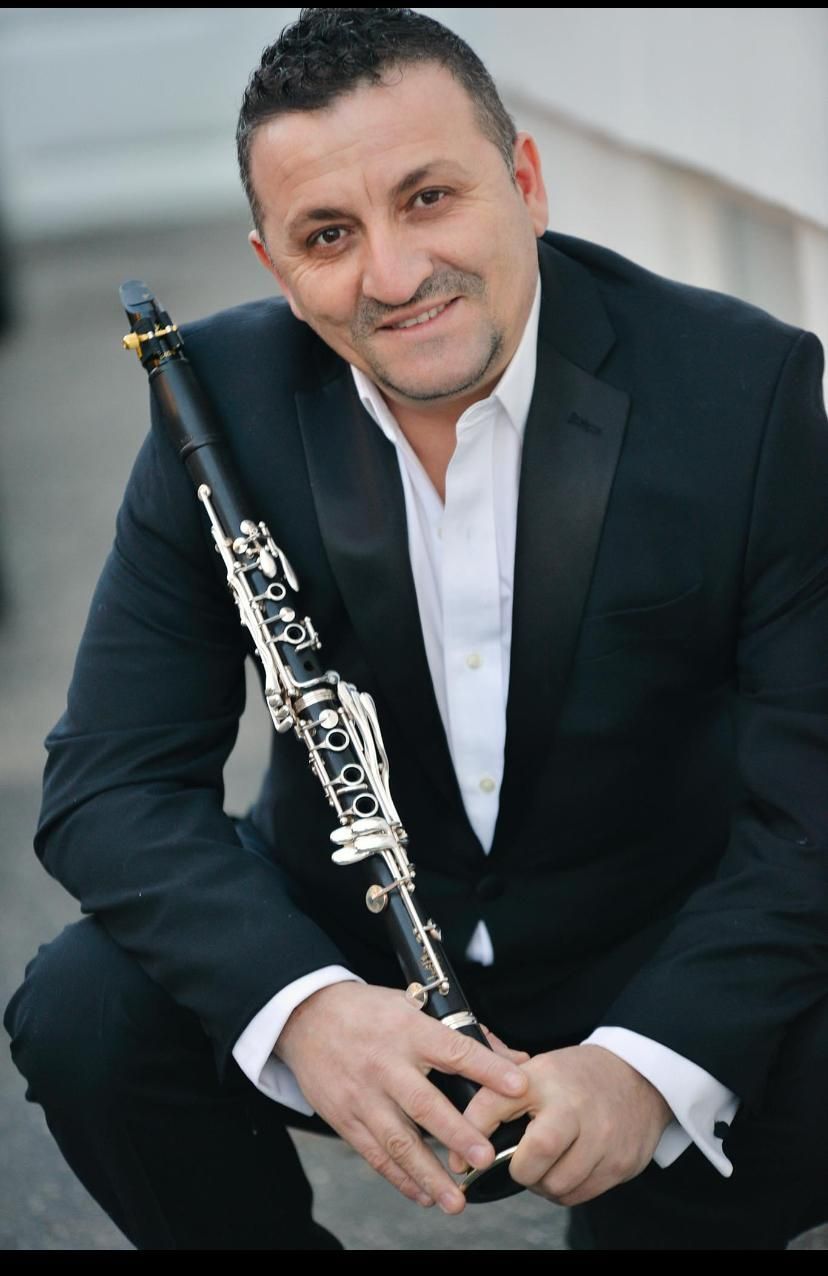 Man in suit smiles, holding a clarinet. Squatting on a balcony or rooftop.