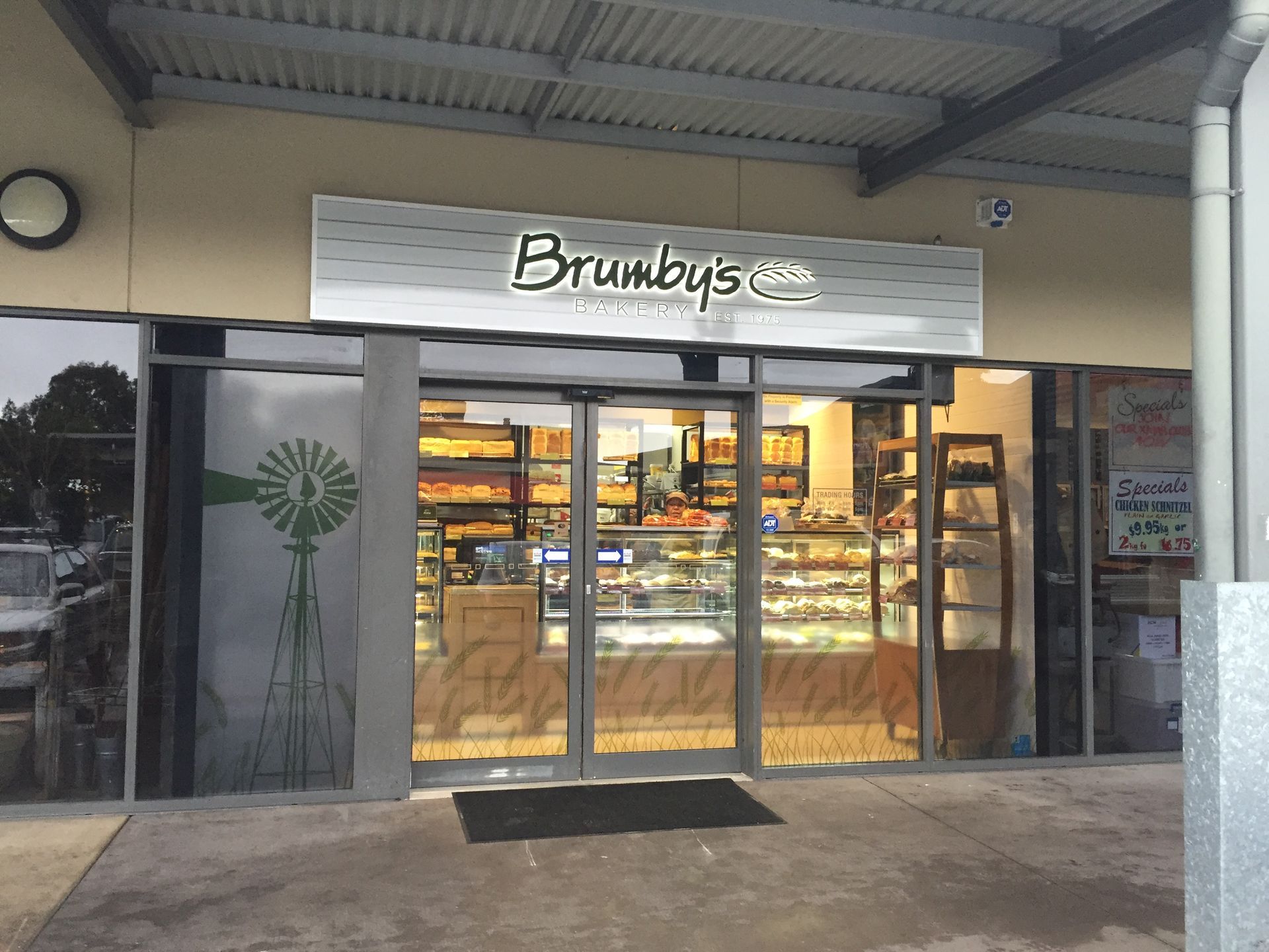 Exterior view of a Brumby’s bakery shop front with glass doors, a silver sign, and a windmill decal on the window.