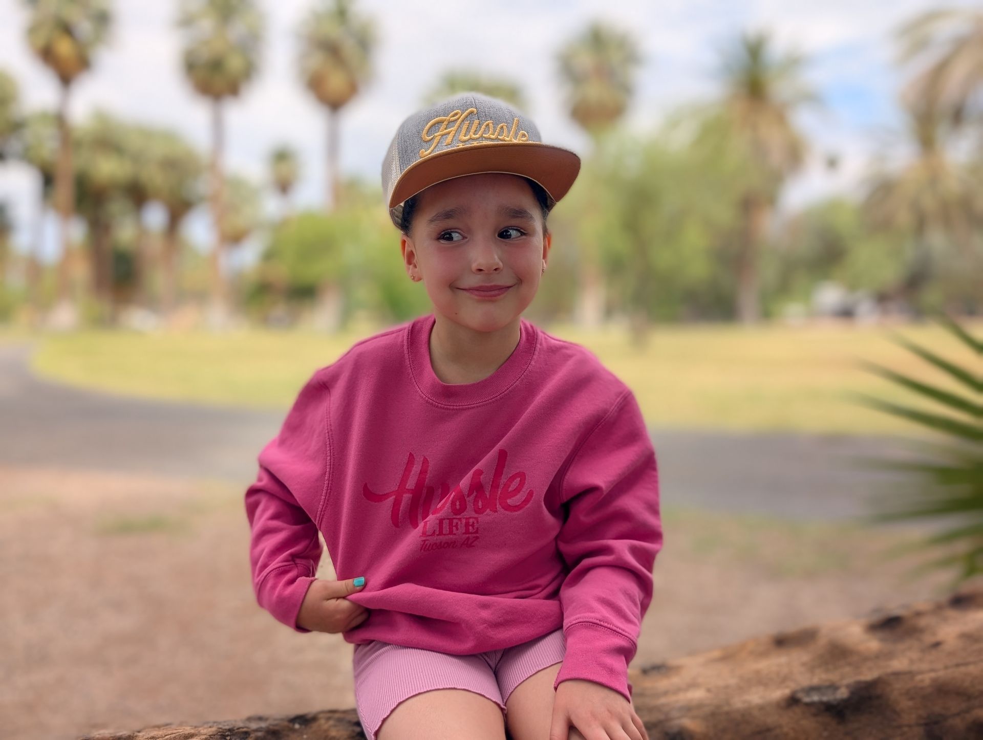 A little girl wearing a pink sweatshirt and a hat is sitting on a log.