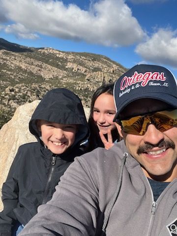 Family selfie on a mountain, smiling. A boy in a black jacket, a girl, and a man with a mustache in a hat and sunglasses.