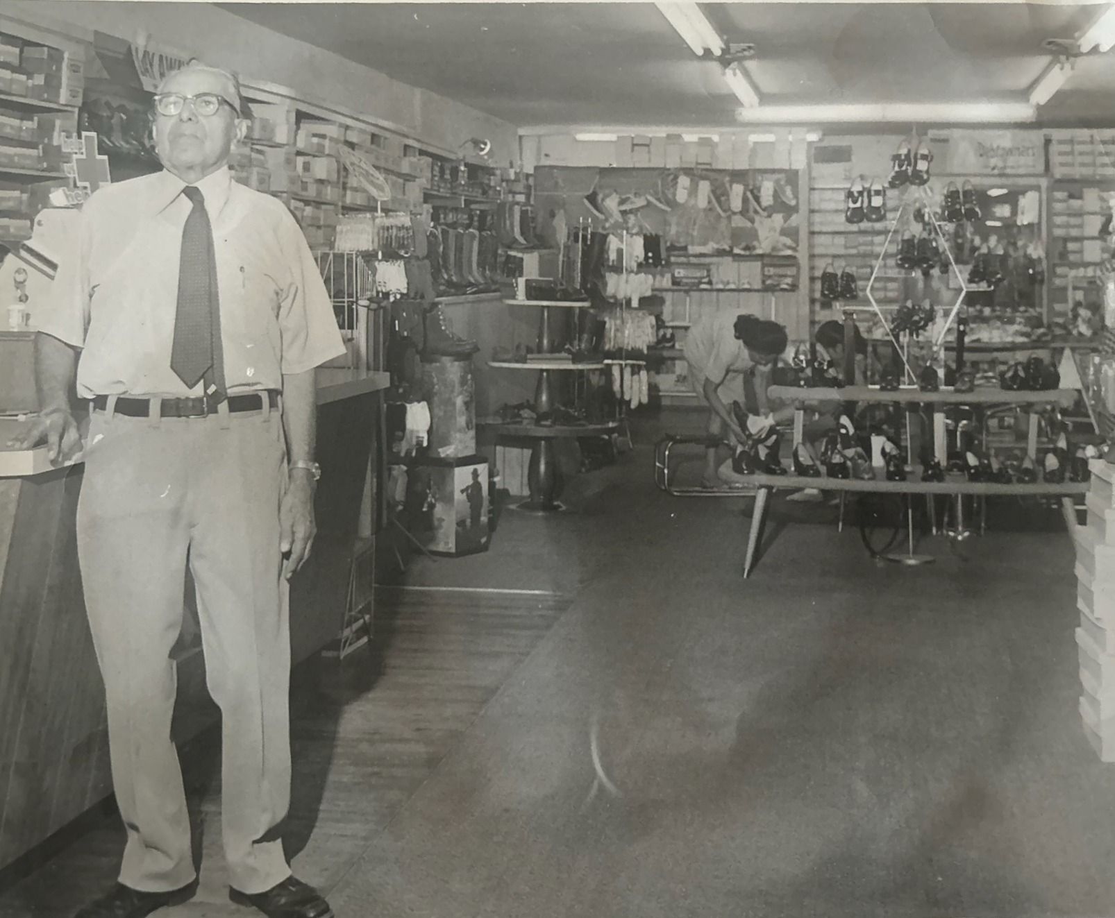 Man in a uniform standing inside a store filled with merchandise on shelves. Black and white photograph.