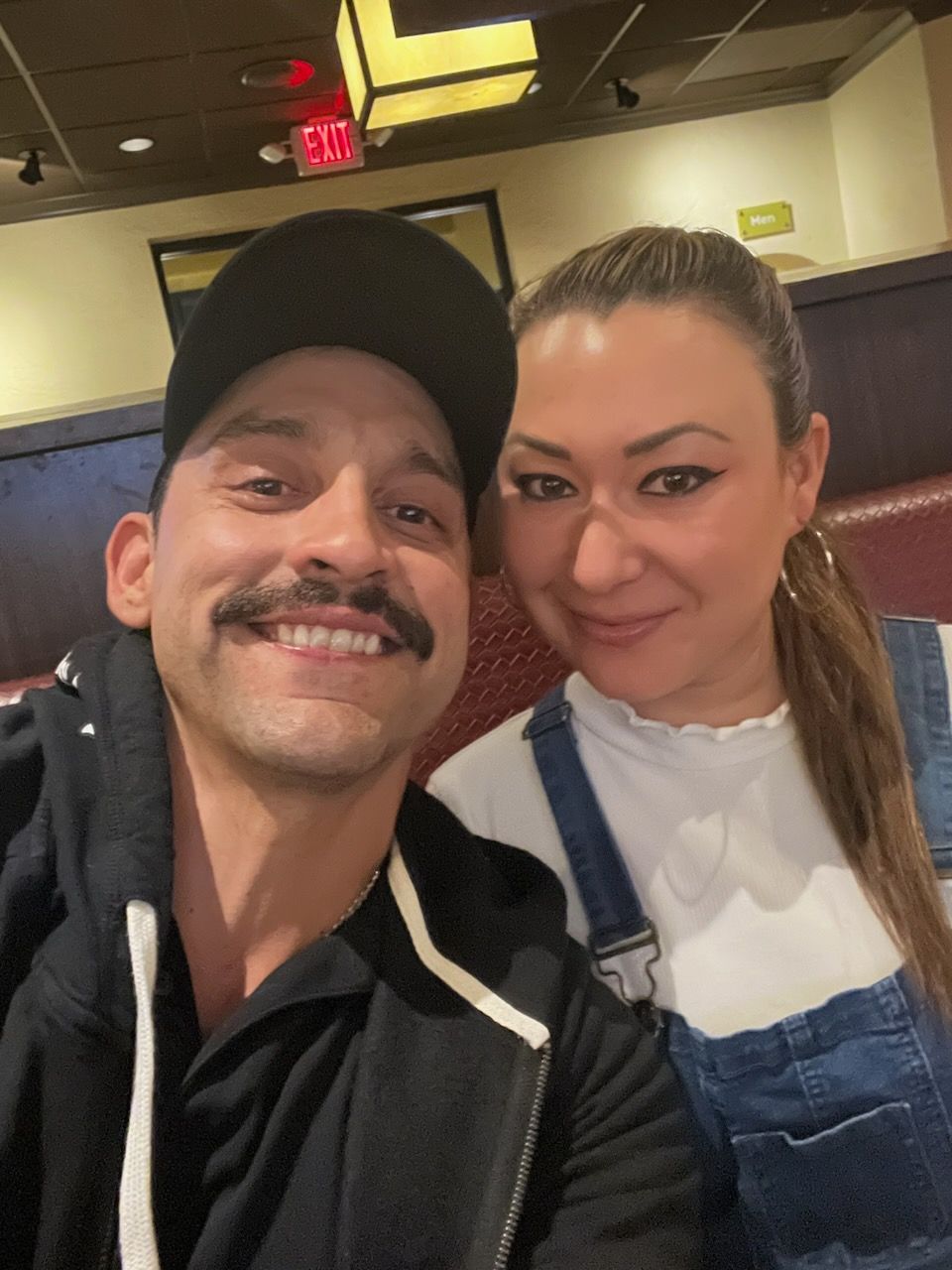 Couple smiling, indoors at a restaurant. Man with mustache, cap. Woman with ponytail, overalls.