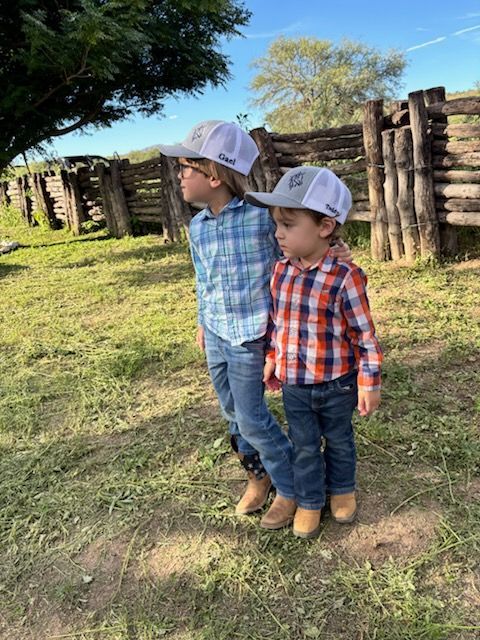 Two children in cowboy hats, shirts, and boots, standing outdoors, looking to the side. Wooden fence in background.