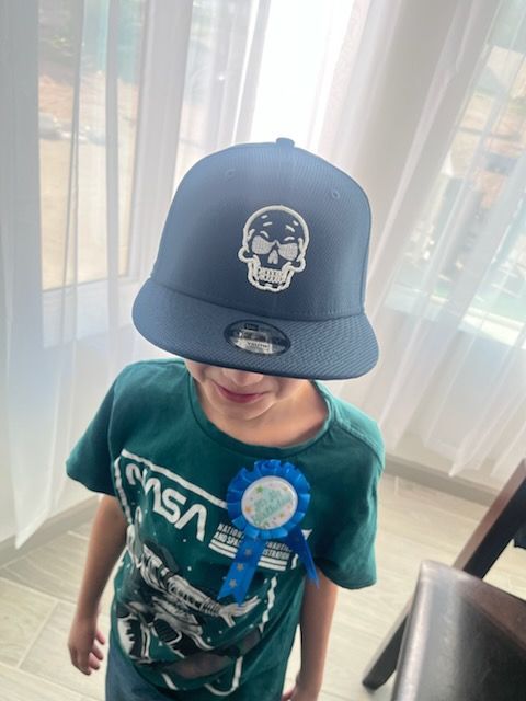 Child wearing a dark blue skull cap and a blue birthday ribbon, indoors.