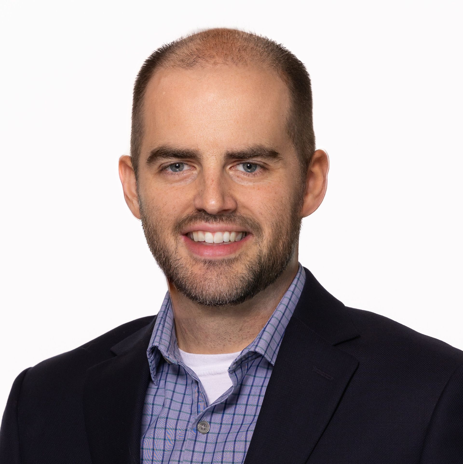A person with short hair and facial hair, wearing a patterned collared shirt and a dark blazer against a white background.