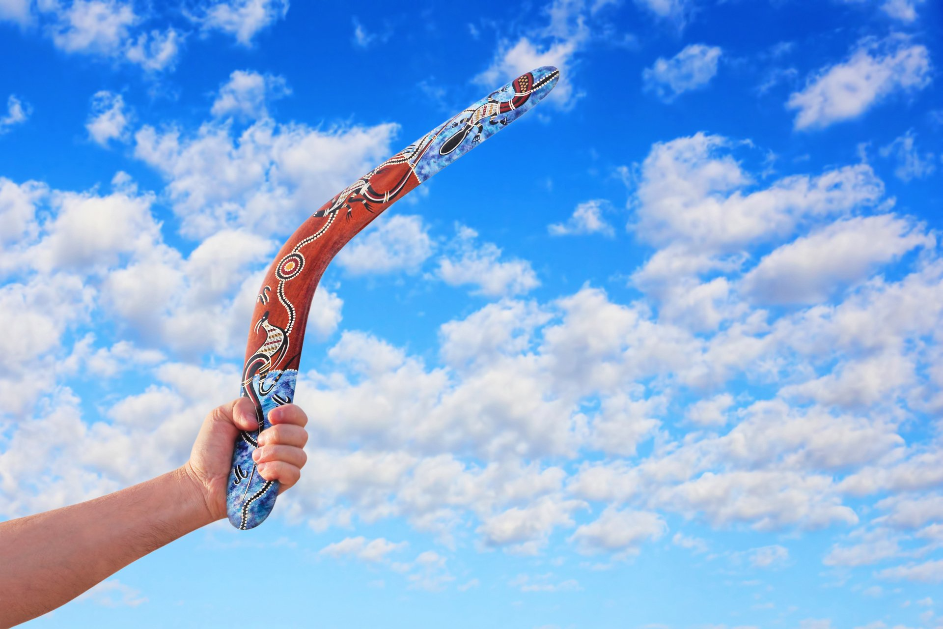 A bright blue sky dotted with clouds in the background, a hand holding a boomerang in the foreground