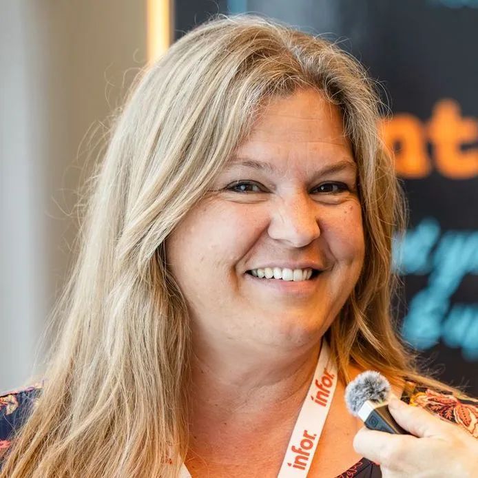 A blonde woman with shoulder length hair smiles at the camera while speaking in a hand-held microphone.