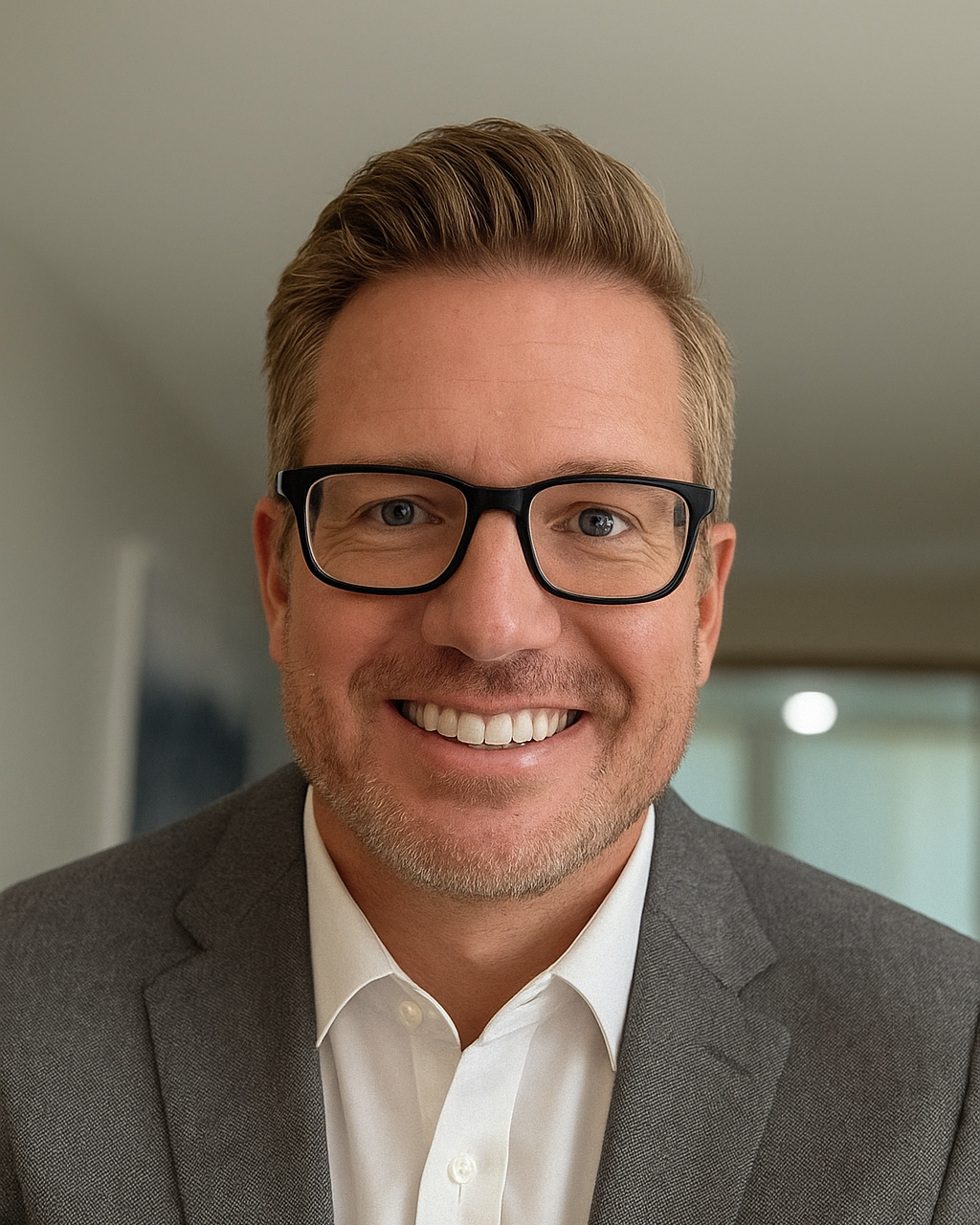 A man with short hair, wearing glasses, a white shirt, and a gray suit smiles at the camera.