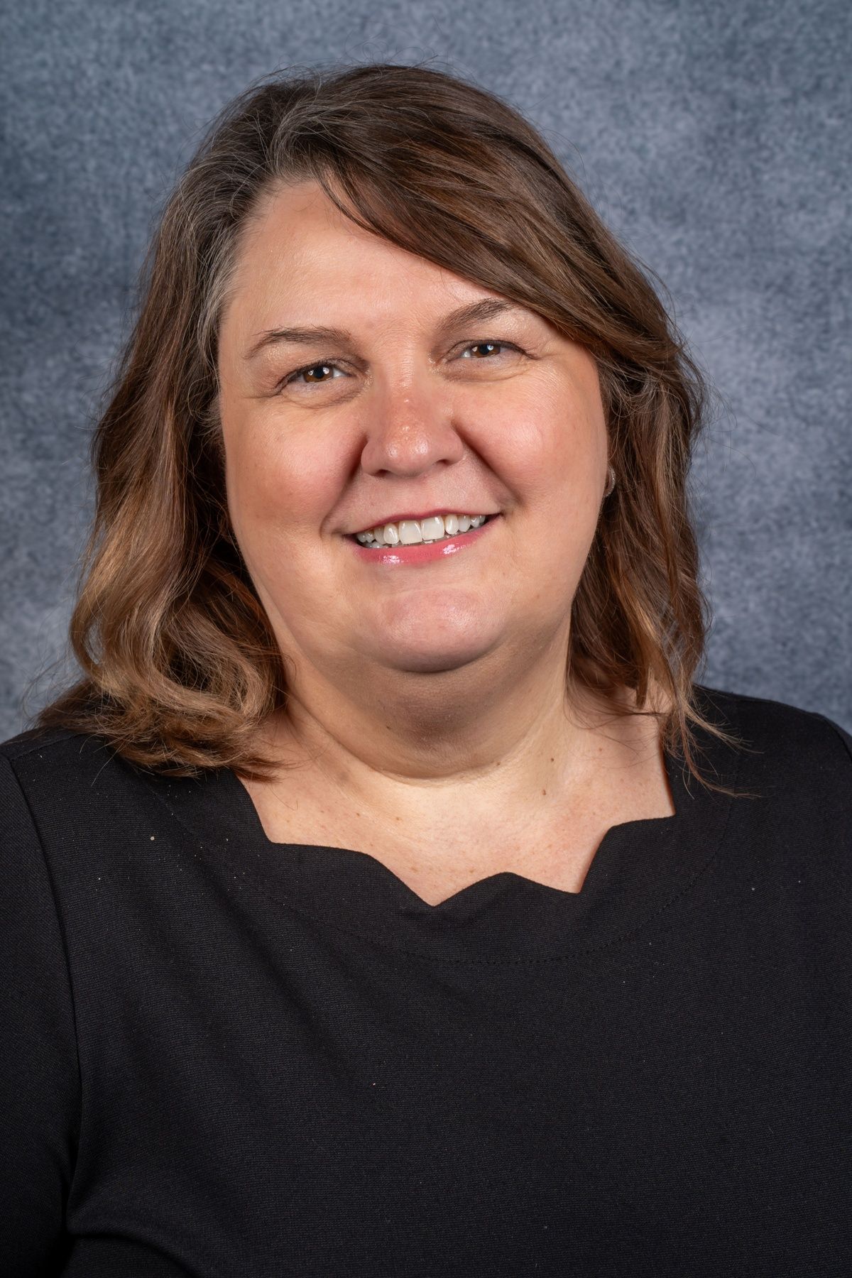 A woman with brown shoulder-length hair, wearing a black top, smiles at the camera against a mottled blue backdrop.