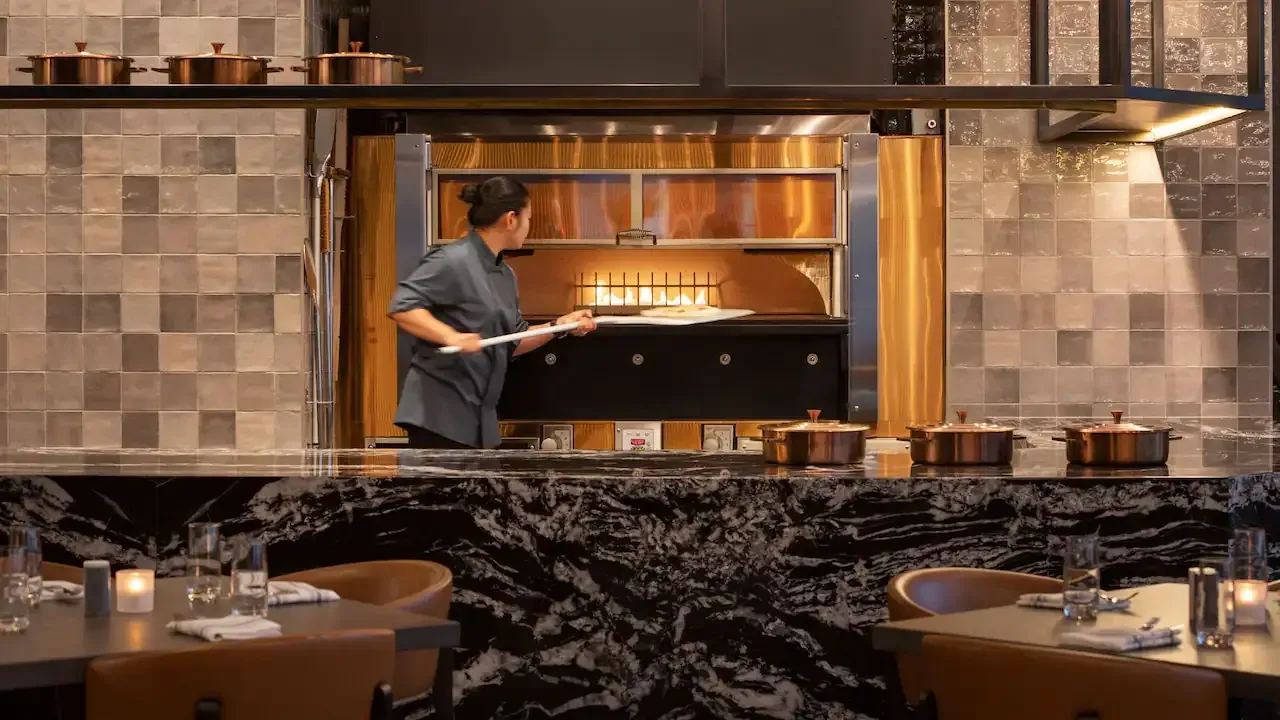 A chef in a grey shirt uses a wooden paddle to pull food out of wood burning oven in a restaurant kitchen.