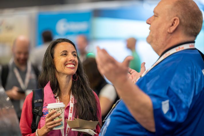 A woman smiles, holding coffee, looking up at a man gesturing with his hands. They both wear conference lanyards.
