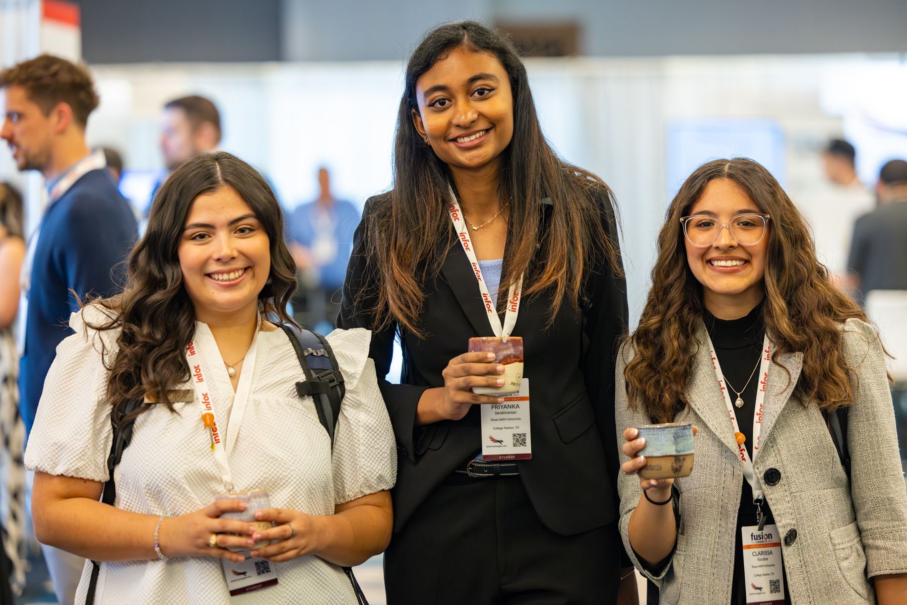 Three young women smiling at a conference. They wear conference badges and business attire. The background is blurred.