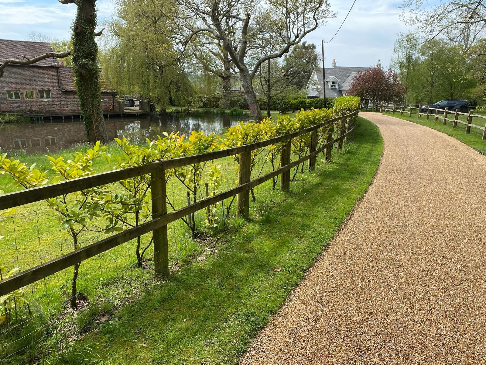 A gravel path curves along a fence and a pond. Lush green grass and trees surround.