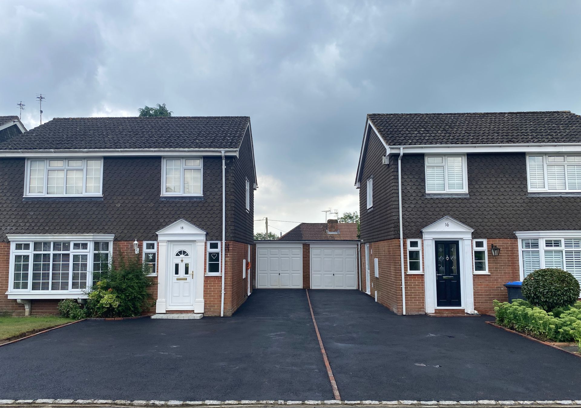 Two nearly identical brick houses with black asphalt driveways and a shared garage under an overcast sky.