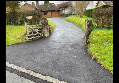 Asphalt driveway curving toward a house, flanked by hedges and grass. Wooden gate on the left.