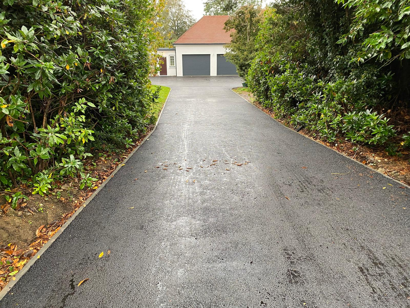 A tarmac driveway leading to a garage surrounded by trees and bushes.