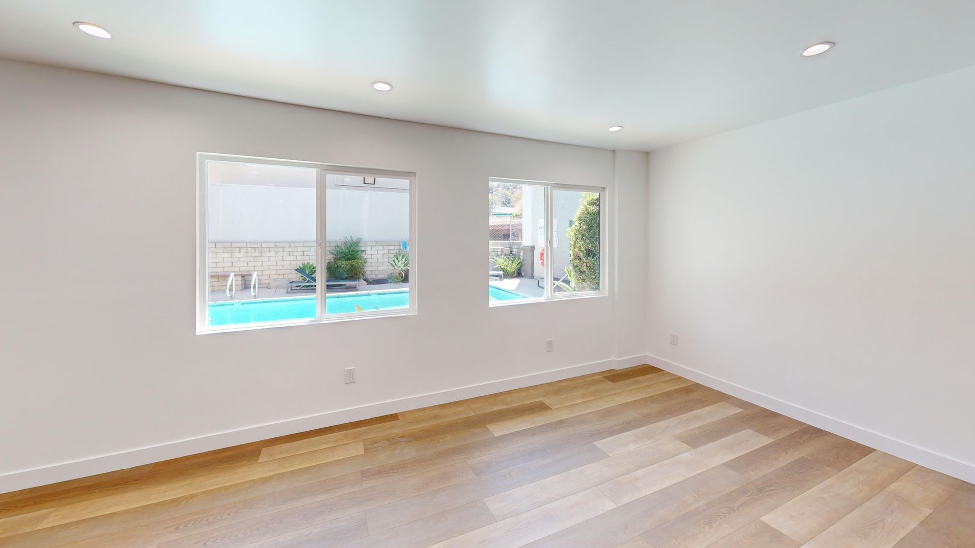 Empty room with hardwood floors, two windows overlooking a pool. Bright white walls, recessed ceiling lights.