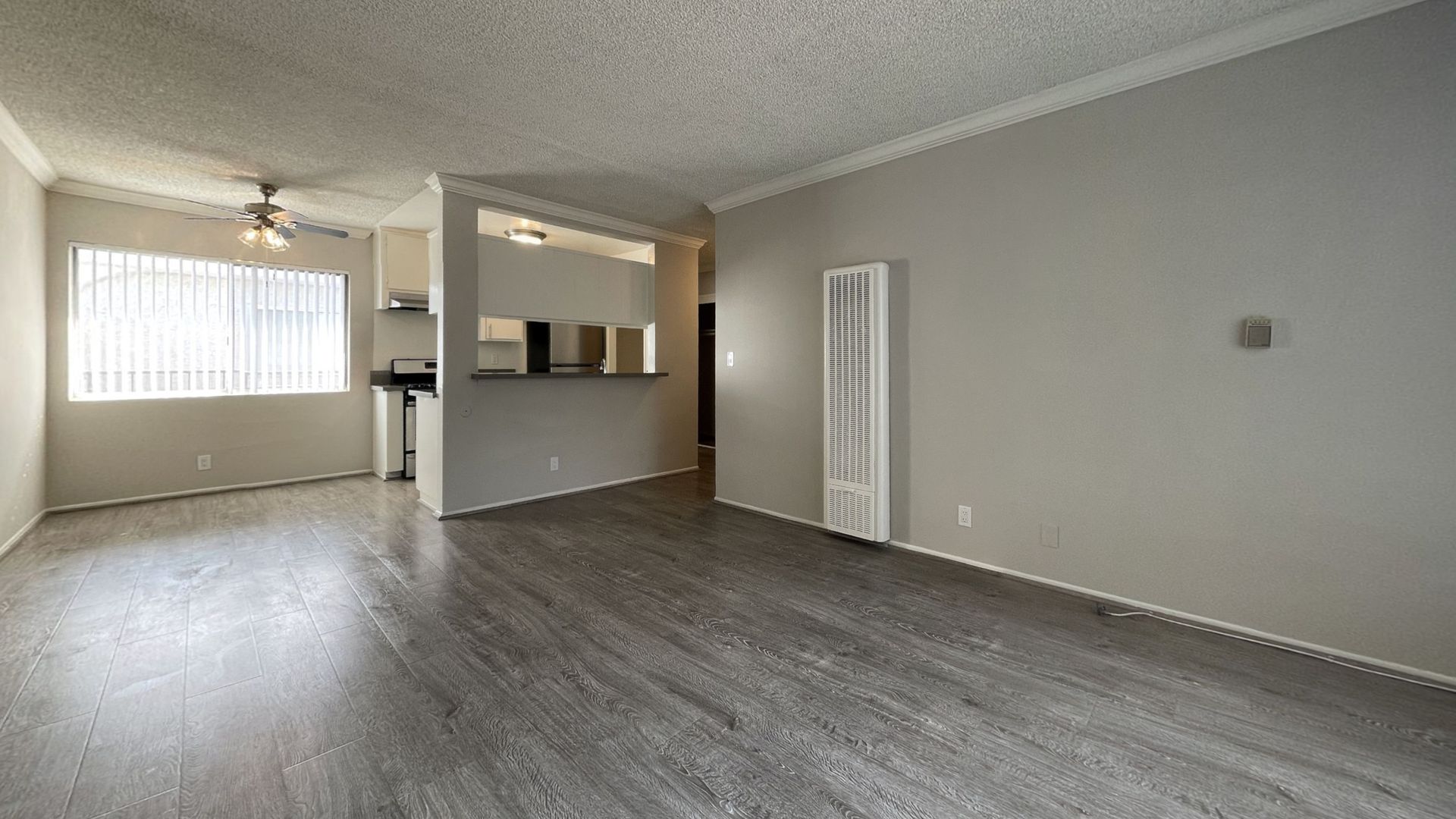 Empty apartment living room with gray flooring, light walls, and a view of the kitchen area.