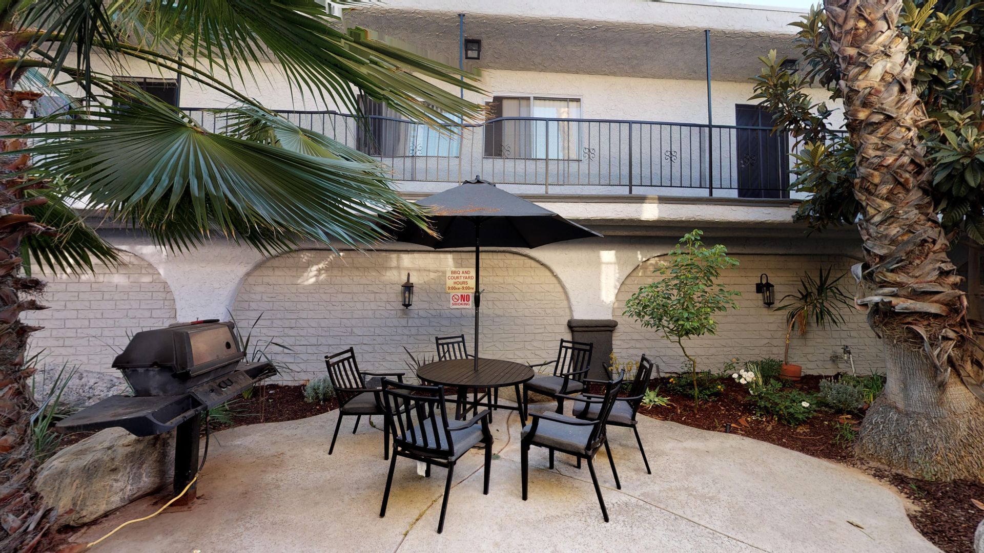 Patio with a black umbrella, table, and chairs. A grill, trees, and a building are visible.
