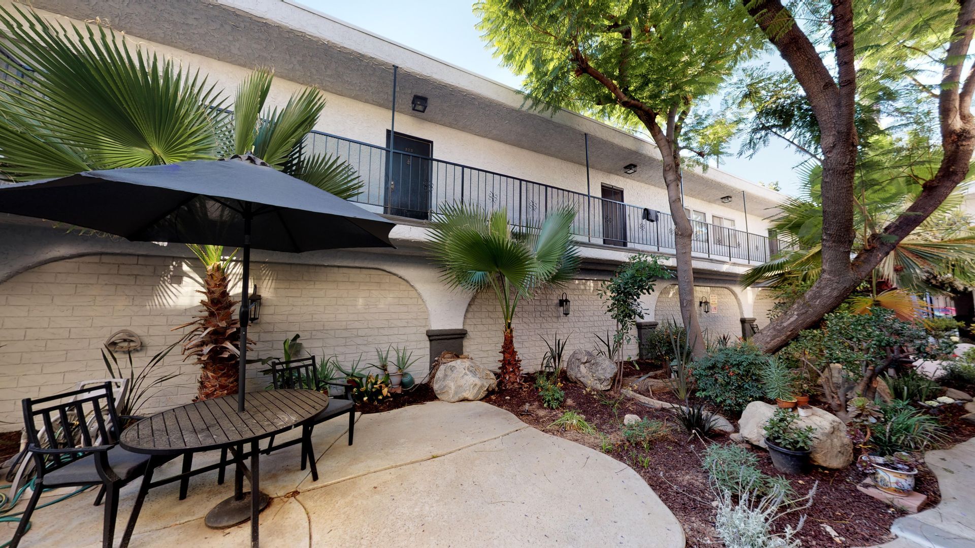 Outdoor patio with black umbrella, table, chairs, and landscaped garden next to a two-story building.