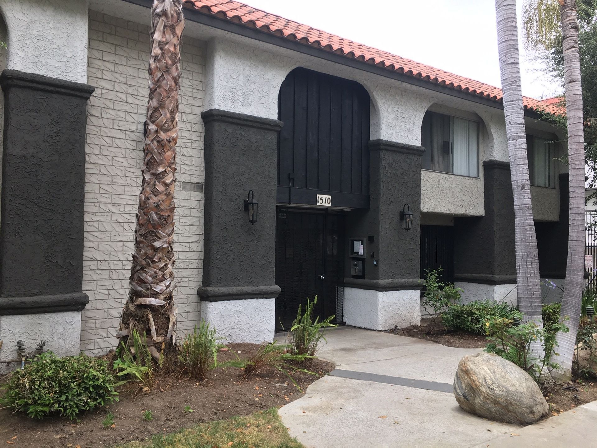 Apartment building with dark entrance, white accents, and red tile roof. Palm tree and rock in foreground.