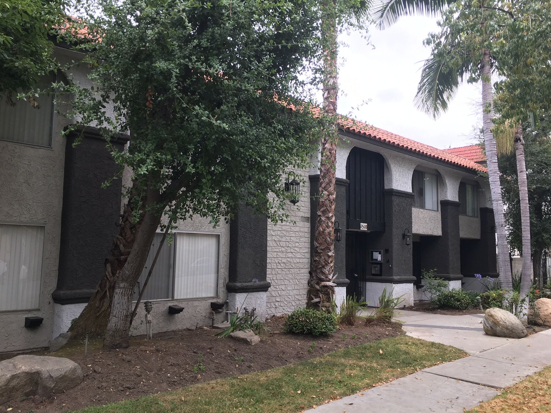 Two-story apartment building with stucco exterior, dark accents, and red tile roof, flanked by trees and grass.