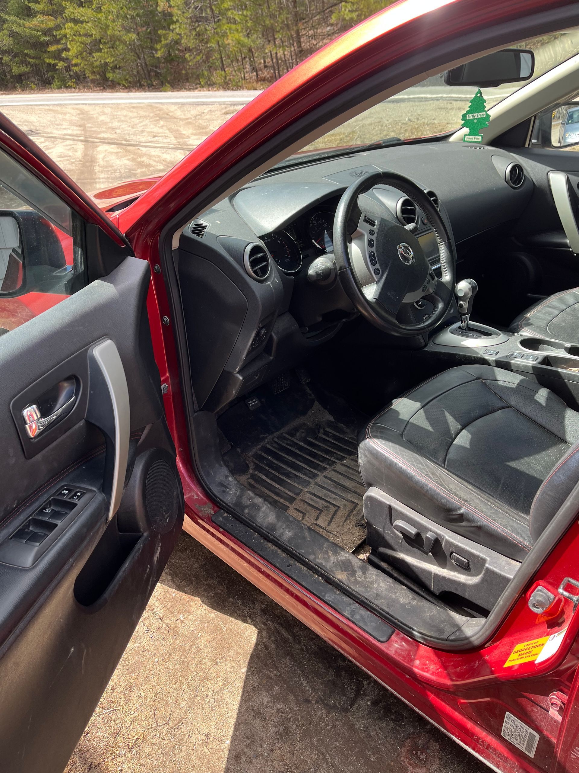 Open red car door showing black interior and steering wheel on a driveway.