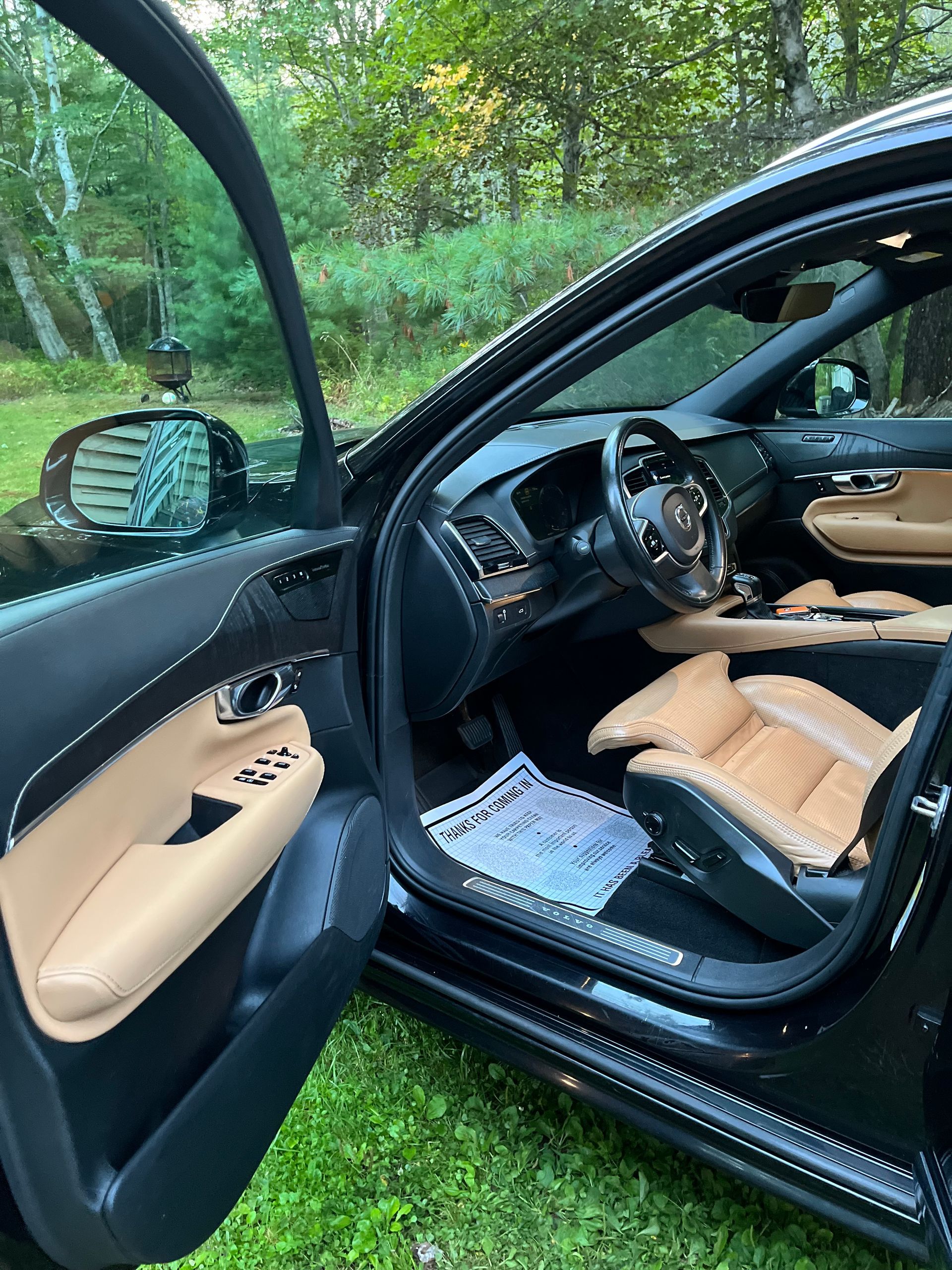 Open car door revealing tan leather interior and steering wheel, parked on grass under trees