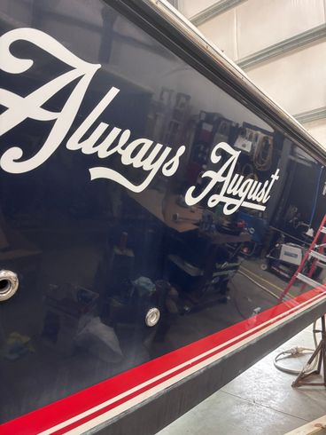 Blue boat hull with white “Always August” lettering and a red stripe in a workshop.