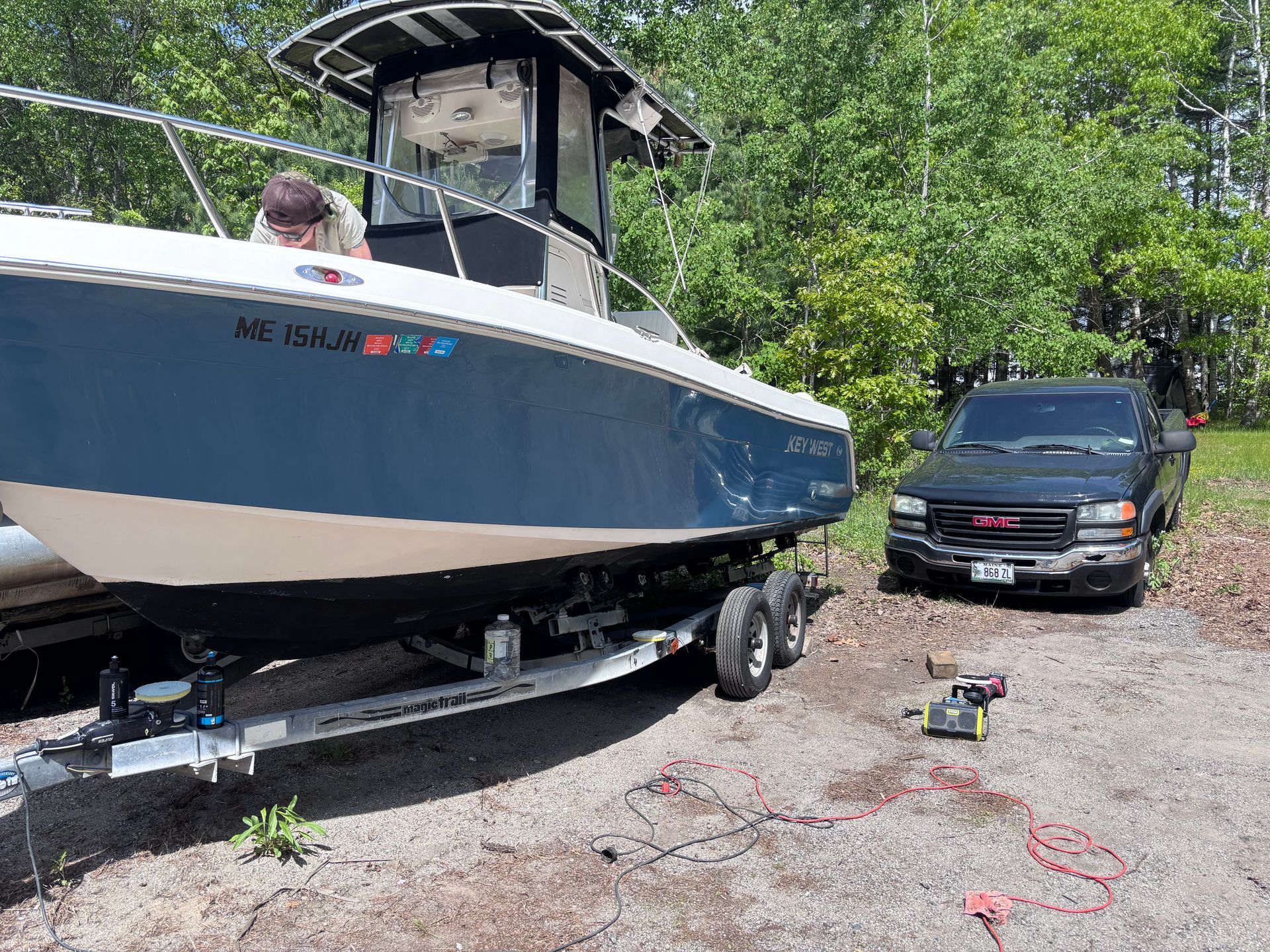 Blue-and-white boat on a trailer beside a black SUV in a wooded driveway, with a person aboard