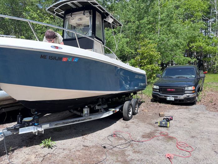 Blue and white boat on trailer in a driveway beside a black SUV and trees