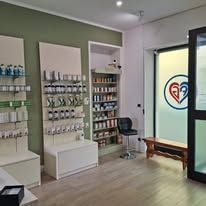 Interno di una farmacia con scaffali di prodotti medicali, un bancone espositivo bianco, una sedia e un logo a forma di cuore sulla porta.