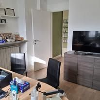 Un ufficio domestico con una scrivania in legno in primo piano, due sedie nere, una porta bianca e un televisore su un mobile scuro.