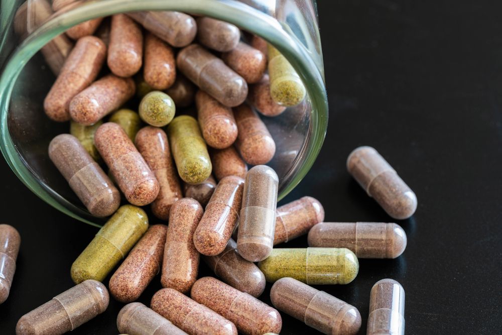Un barattolo di vetro si è rovesciato, spargendo capsule di integratori a base di erbe di vari colori, marroni e verdi, su una superficie nera.