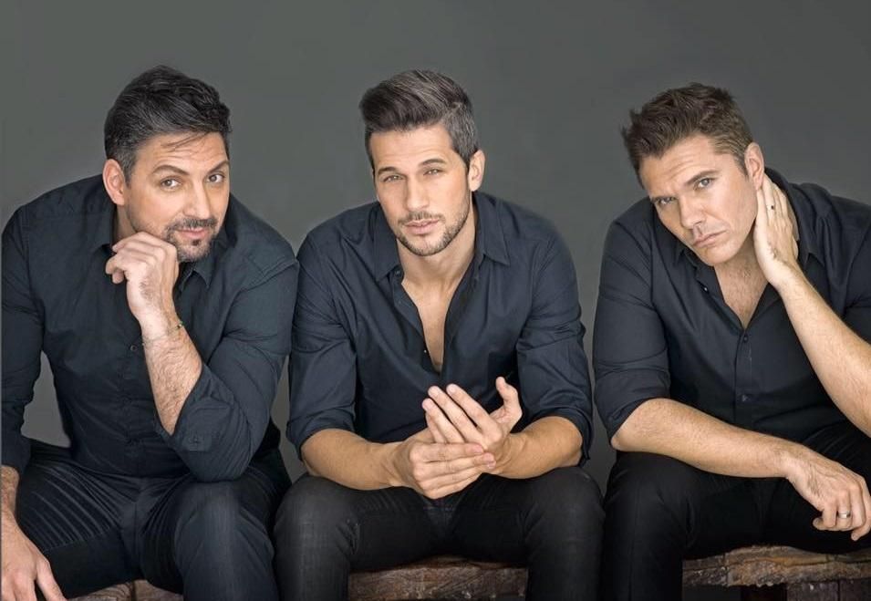 Tres hombres con camisas negras están sentados uno al lado del otro en taburetes de madera.