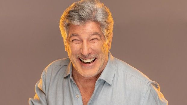 Un hombre con cabello gris y barba sonríe y hace una cara divertida.