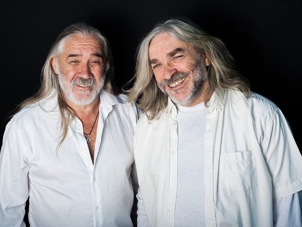 Dos hombres con pelo largo y barba están parados uno al lado del otro y sonríen.