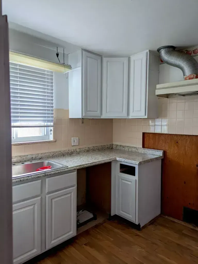 White kitchen cabinets and countertops in a corner space. A window with blinds, exhaust vent, and hardwood floor are visible.